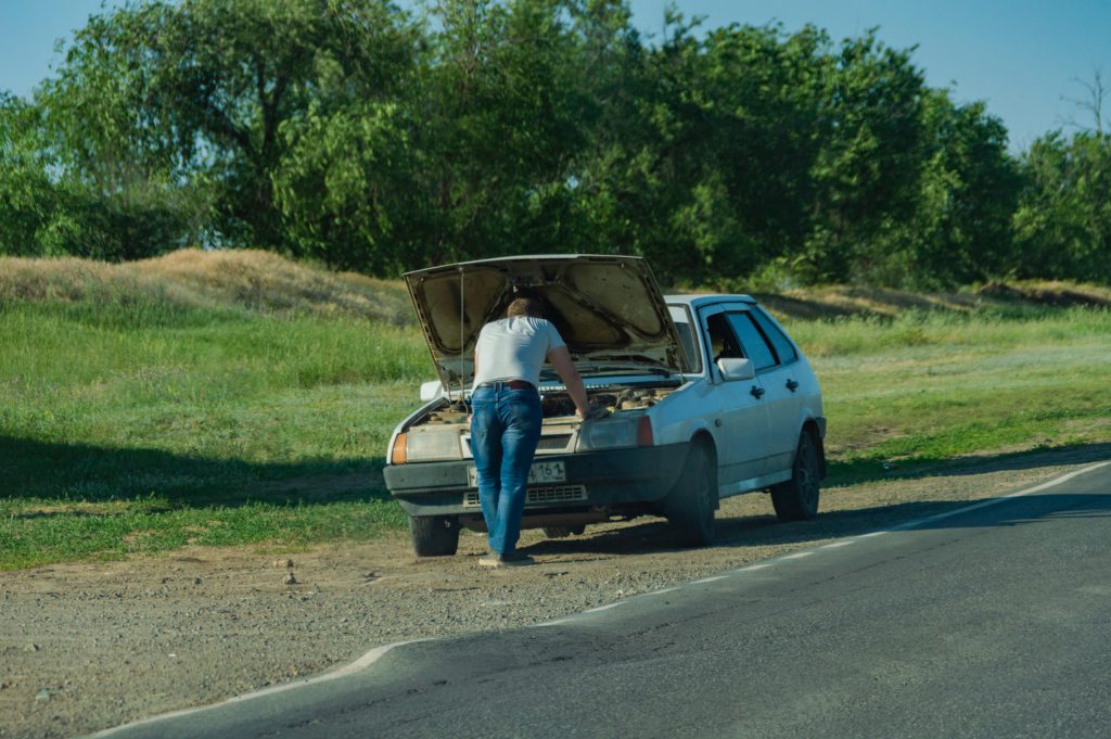 A man stands by his car on the roadside, inspecting a breakdown issue.