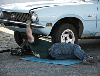 A mechanic working under a classic car on the pavement during the day.