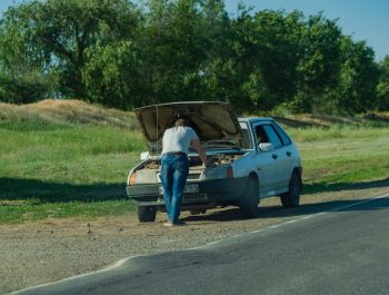 A man stands by his car on the roadside, inspecting a breakdown issue.